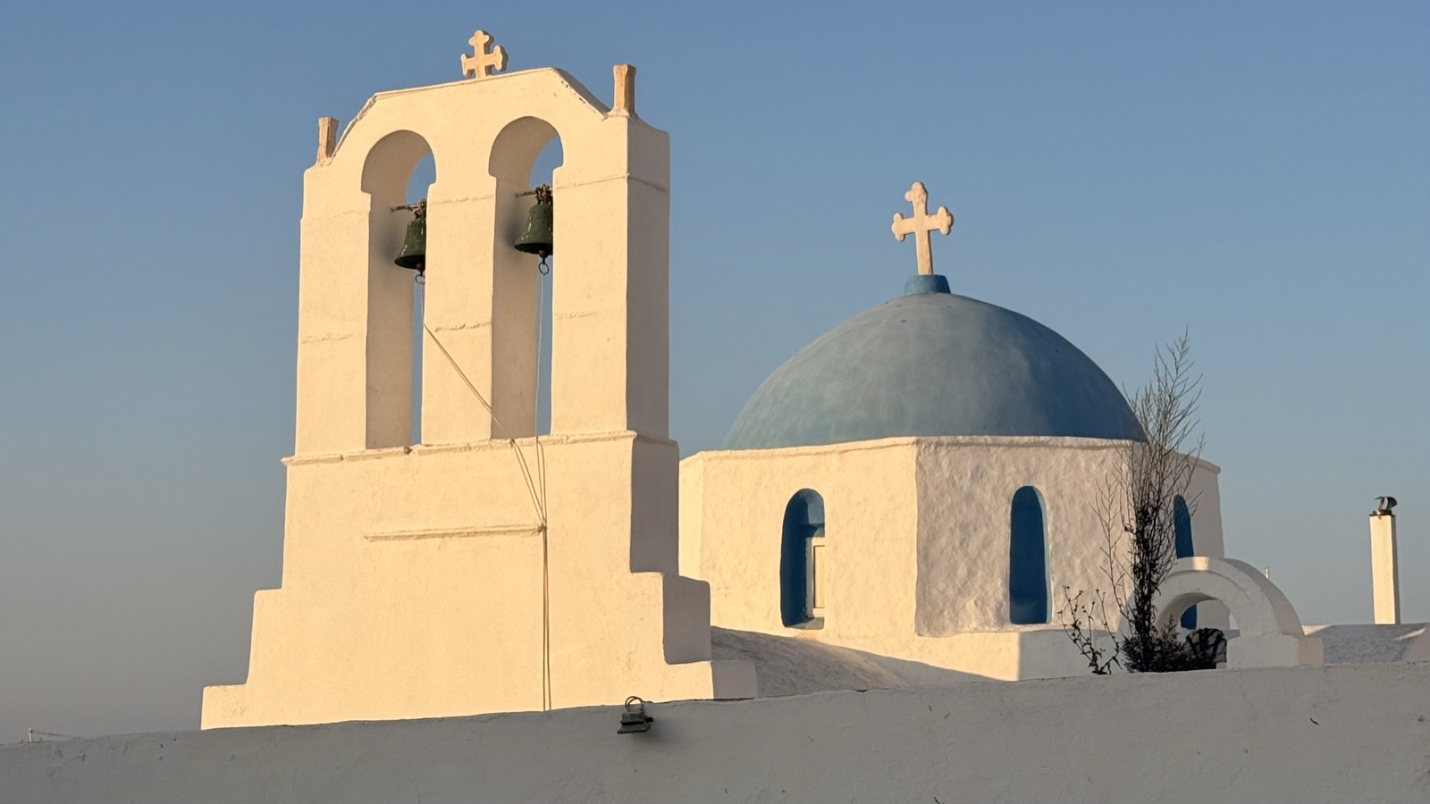 A white Greek church with a blue dome and bell tower at golden hour, on the island of Paros